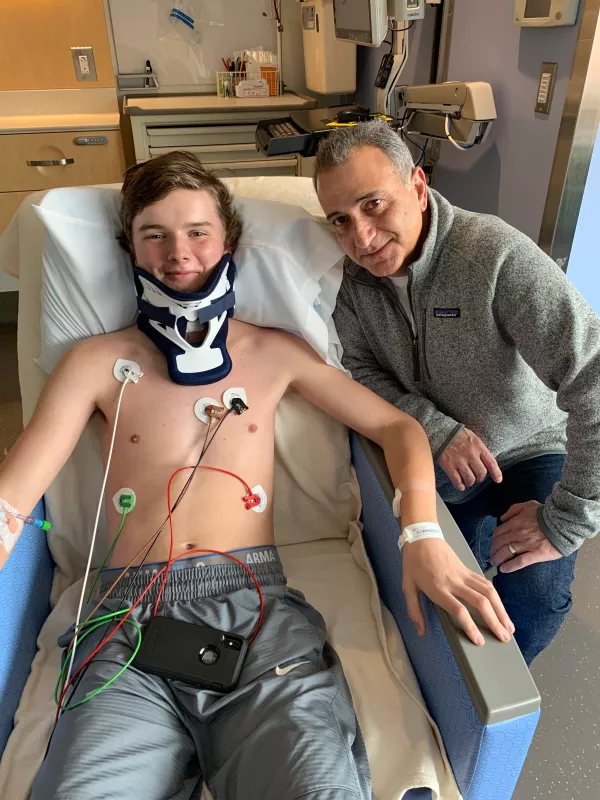 Jackson Webber in a neckbrace, laying in a hospital bed, with Dr. Cassim Igram next to him