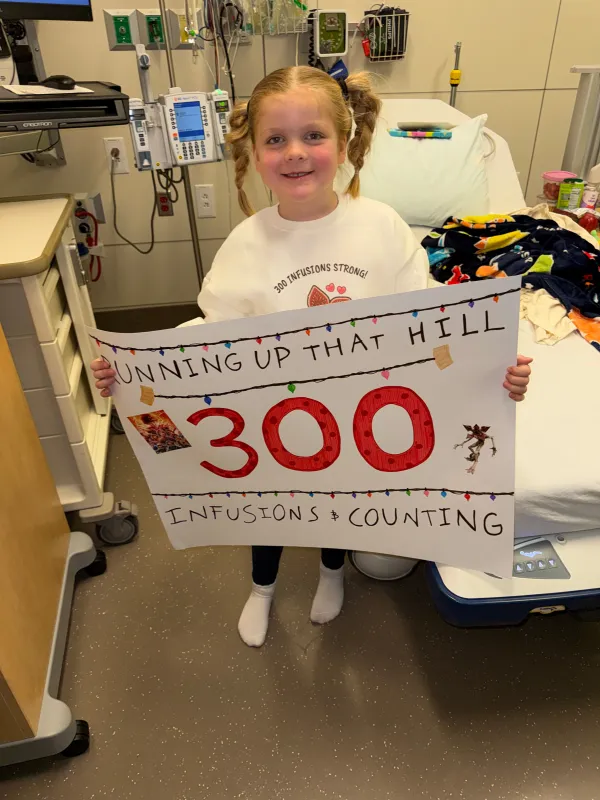 Kid Captain Harlow Taube in the hospital holding a sign announcing her 300th infusion