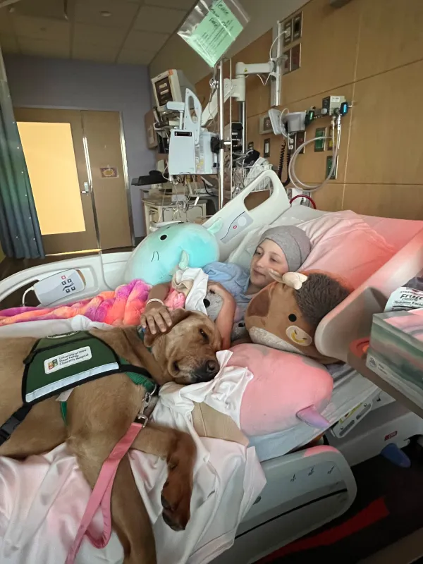 Kid Captain Kaelyn O’Brien laying in a hospital bed with a therapy dog