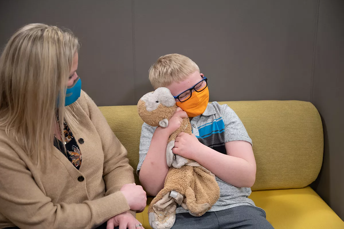 Child hugs a stuffed monkey toy while sitting with their mother in a waiting room